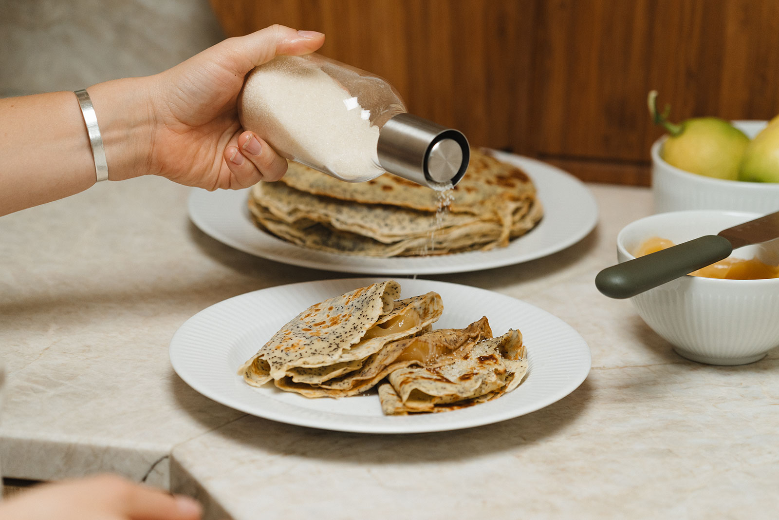 Auf einer Arbeitsfläche sind frisch gebratene Crepes angerichtet auf Legio Nova Frühstückstellern von Eva Trio. Eine Hand streut Zucker auf die dünnen Pfannkuchen.