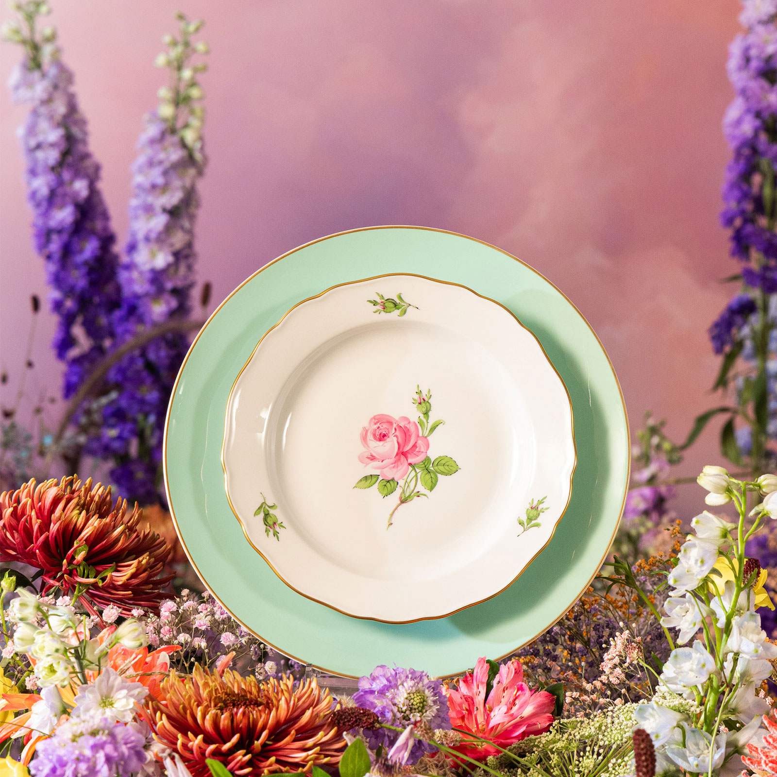 Vor Hintergrund aus arrangierten bunten Blumen steht ein Teller des Dekors Meissener Rose auf dem seladongrünen Urban Colors Speiseteller von Meissen.
