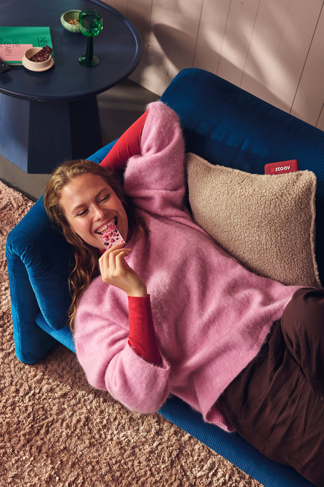 Auf einem blauen Sofa kuschelt eine lächelnde Frau in einem pinken Pulli mit dem Ploov Heizkissen Woolly Beige von Stoov.