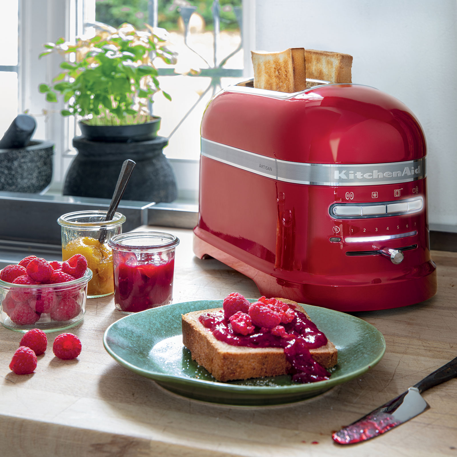 Mit frisch getoastetem Brot in den Schlitzen steht der Artisan Toaster auf der Arbeitsplatte. Im Vordergrund liegt ein reich mit roter Marmelade bestrichenes Brot.