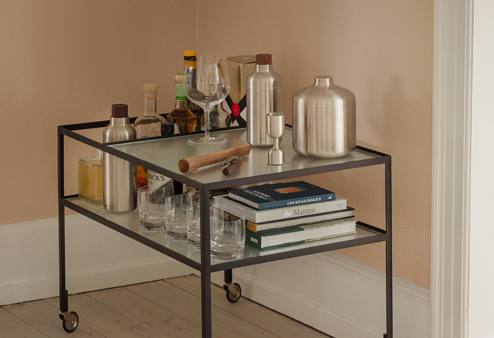 Ein Barwagen vor beiger Wand. Auf dem wagen befinden sich der Cocktailshaker sowie weitere Produkte der Marke Eva Trio.