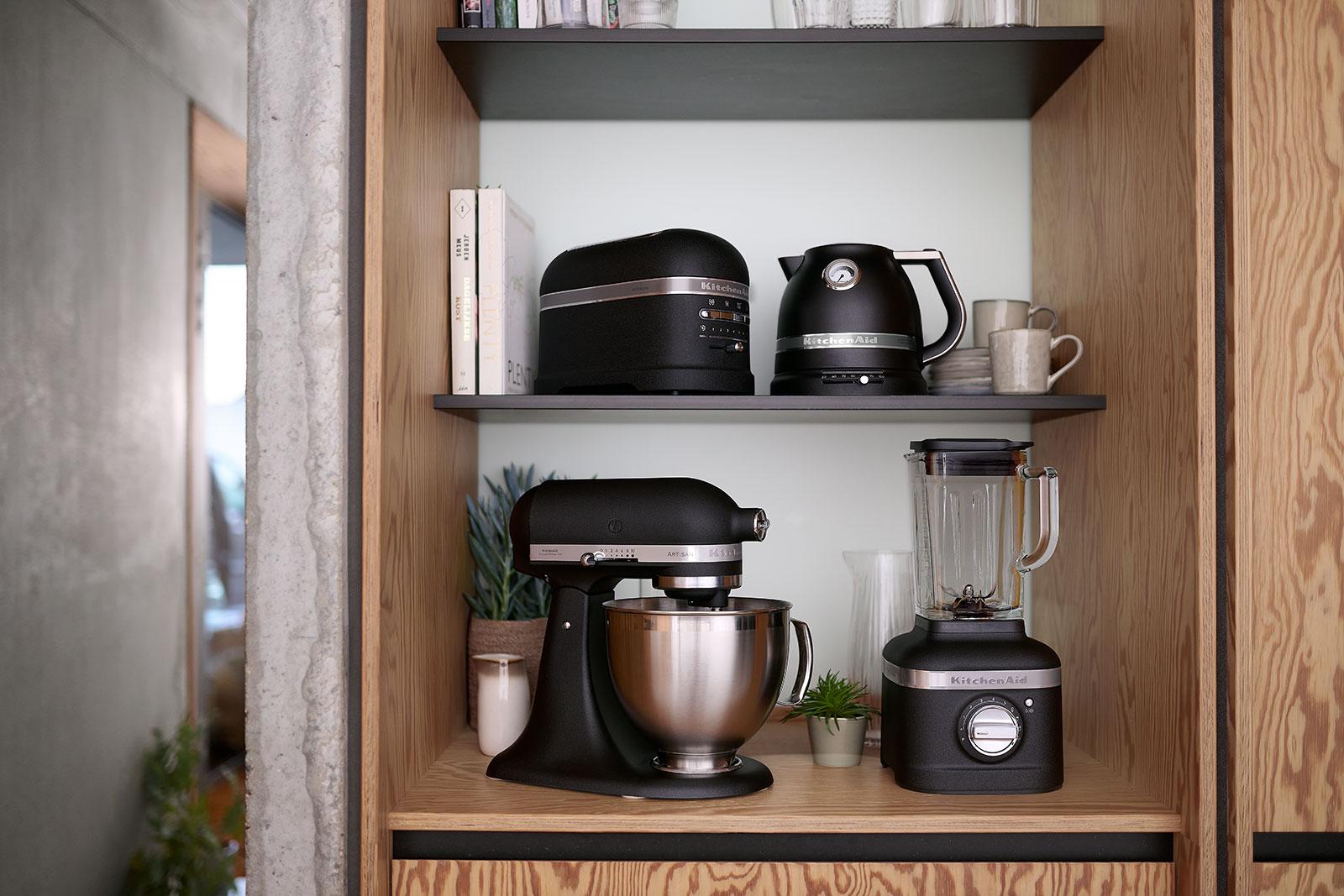 Die Suite der Artisan Artikel von KitchenAid in der Farbe gusseisen schwarz ist in einem Holzregal ausgestellt. Das umfasst den Toaster, den Wasserkocher, die Küchenmaschine und den Standmixer.