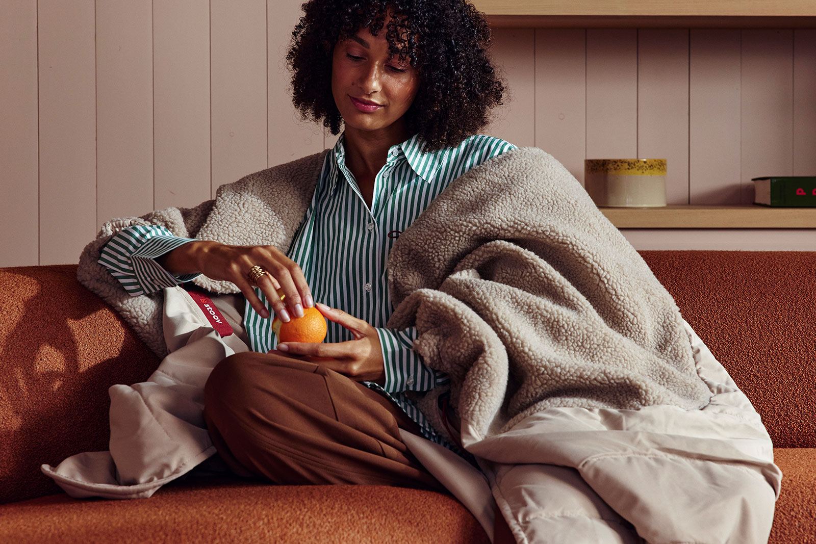 Eine Frau sitzt eingehüllt in die Huge Hug Heizdecke Woolly Soft Beige auf dem Sofa und pellt eine Mandarine.