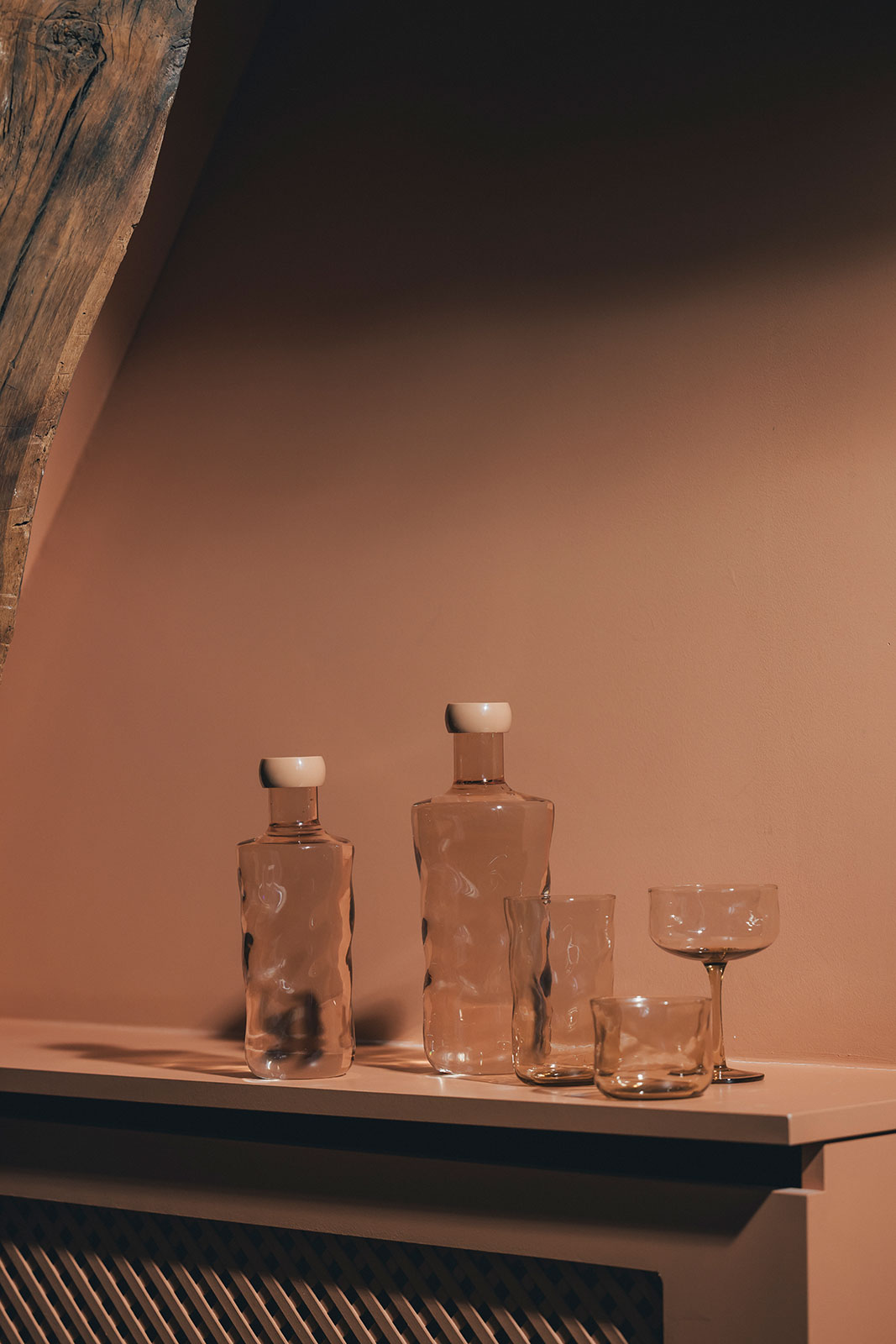 Vor einer terrakottafarbenen Wand stehen mehrere Wasserflaschen und Trinkgläser aus pfirsichfarbenem Glas.