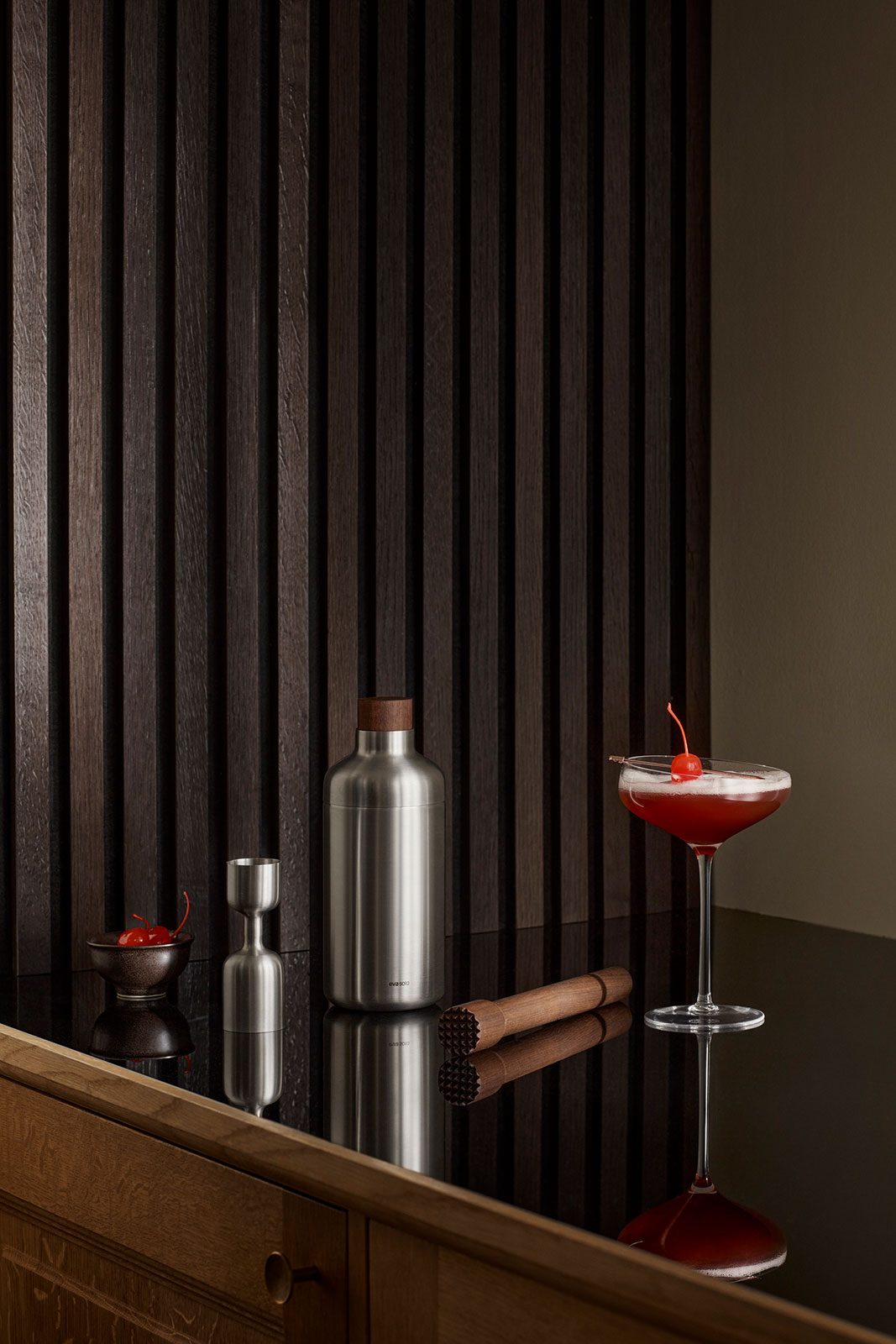 Ein Sideboard in abgedunkelter Atmosphäre, auf dem sich ein Cocktailglas, der Stößel sowie weitere Cocktail Zubereitungsaccessoires befinden.