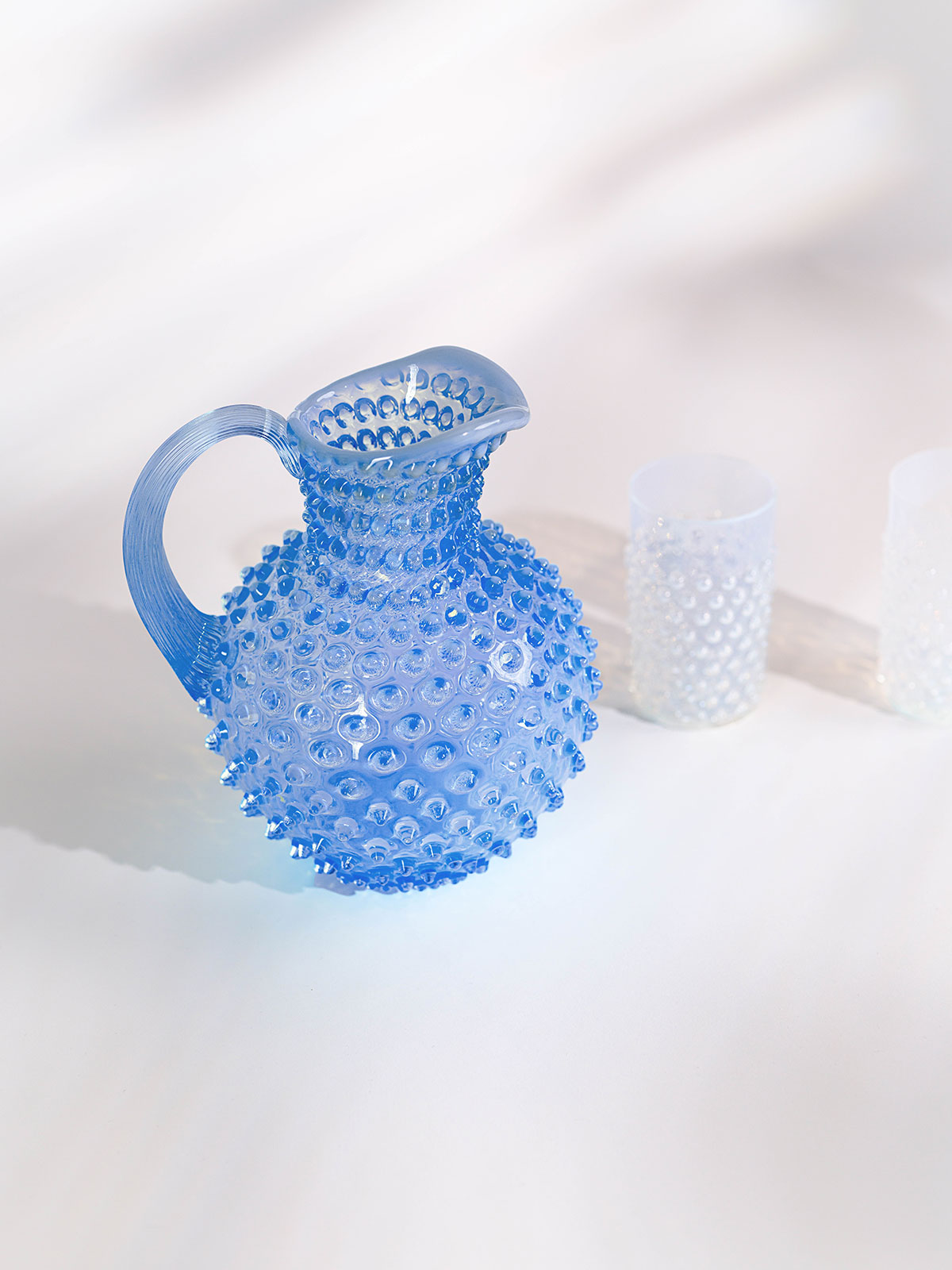 Vir einem weißen Hintergrund steht der Paris Hobnail Krug Light blue von Anna von Lipa neben weißen Tringläsern, um zu zeigen, wie lebhaft seine Farbe ist.