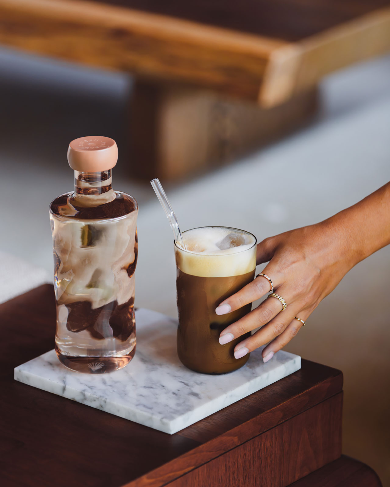Auf einem Mamorstück steht neben einer Wasserflasche der gleichen Farbe ein pfirsichfarbenes Glas mit einem Kaffeegetränk darin. Von rechts greift eine Hand nach dem Glas.