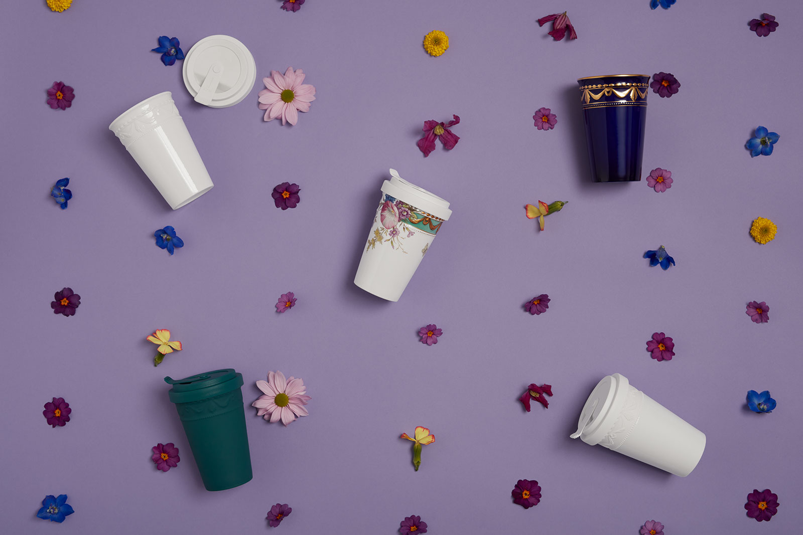 Auf einem fliederfarbenen Untergrund liegen verschiedene KPM To-Go Becher inmitten verstreuter frischer Blumen.