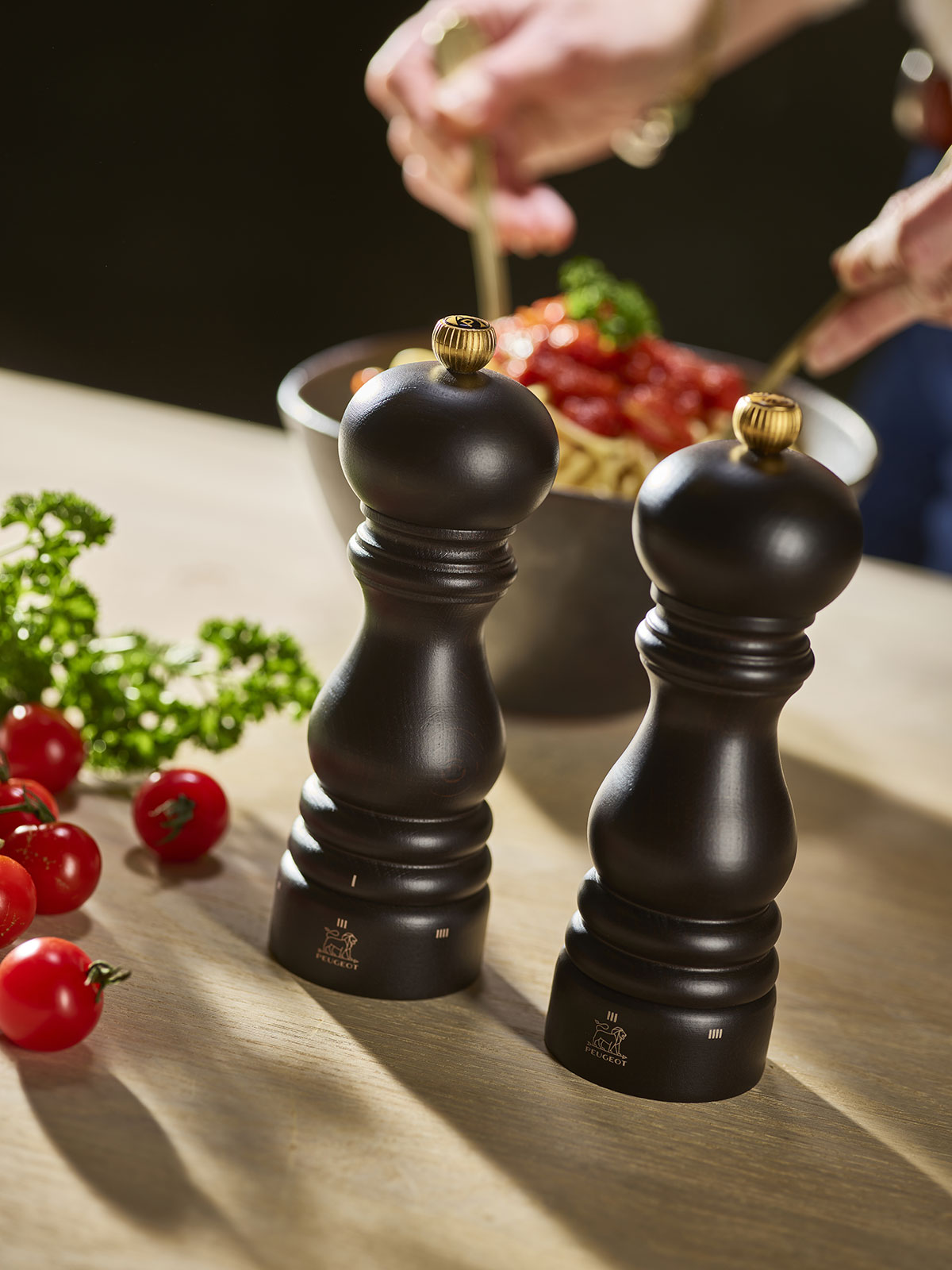 Würzbereit steht ein Paar aus Paris Pfeffer- und Salzmühle in schokolade im Vordergrund, während jemand im Hintergrund jemand Spaghetti anrichtet.