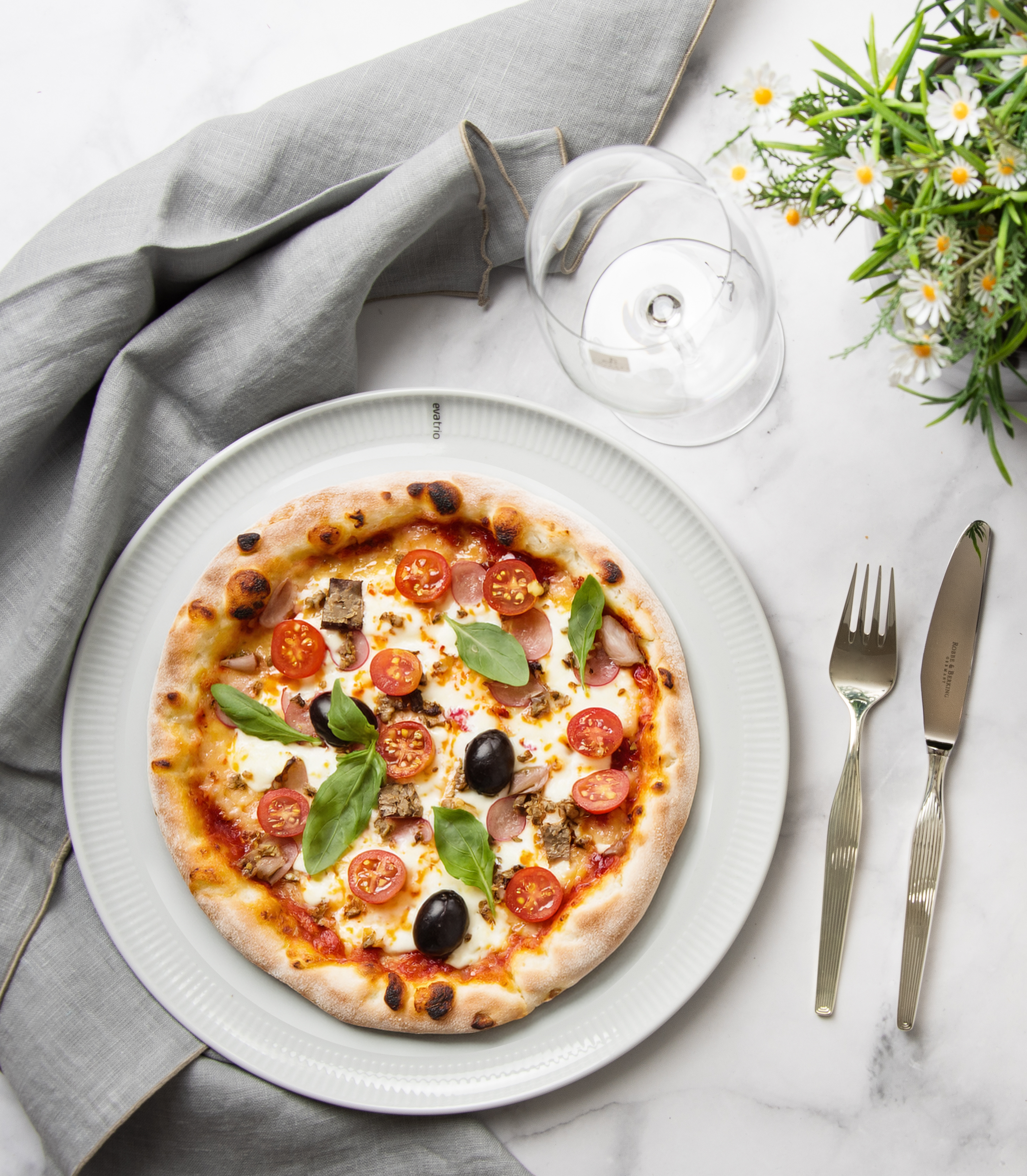Ein gedeckter Marmortisch von oben fotografiert. Auf dem Tisch steht ein Pizzateller, ein Weinglas, Besteck sowie Blumen und das hellgraue Geschirrtuch von Erika Vaitkute.