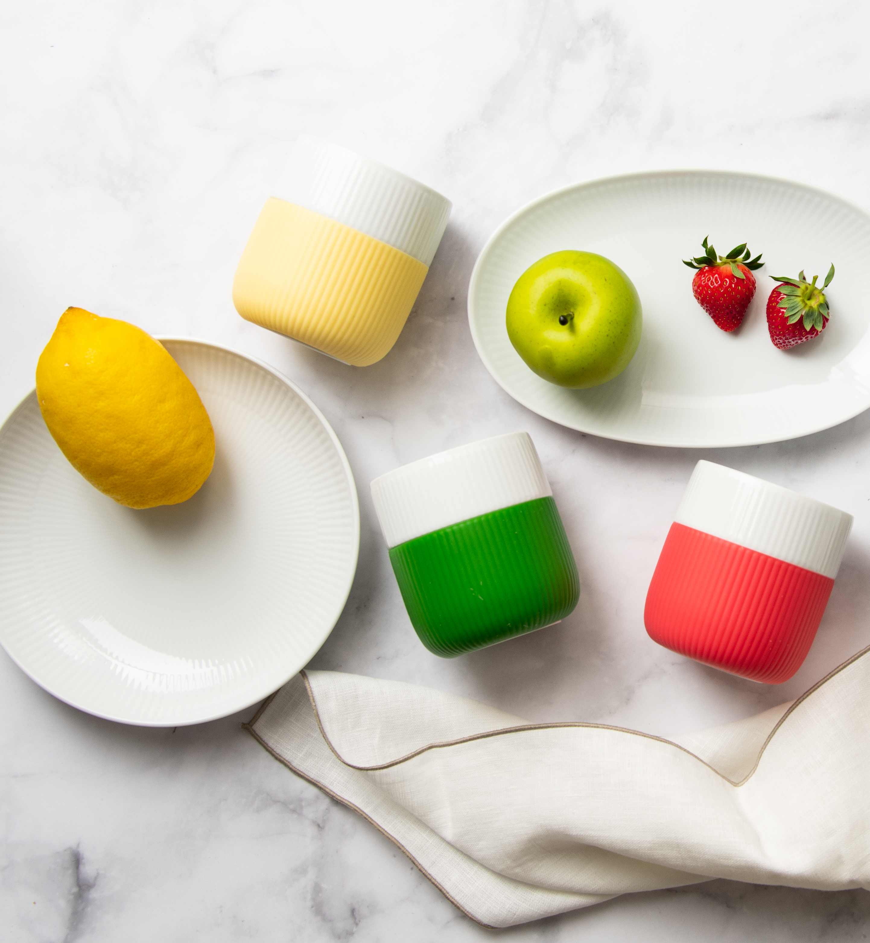 Ein gedeckter Marmortisch von oben fotografiert. Auf dem Tisch stehen weißes Geschirr, bunte Becher und darunter liegt das weiße Erika Vaitkute Geschirrtuch.