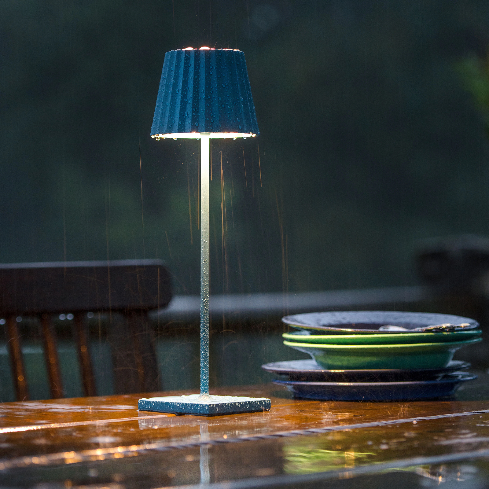 Eine blaue Akku-Tischleuchte auf einem dekorierten Outdoortisch, die im regen steht.