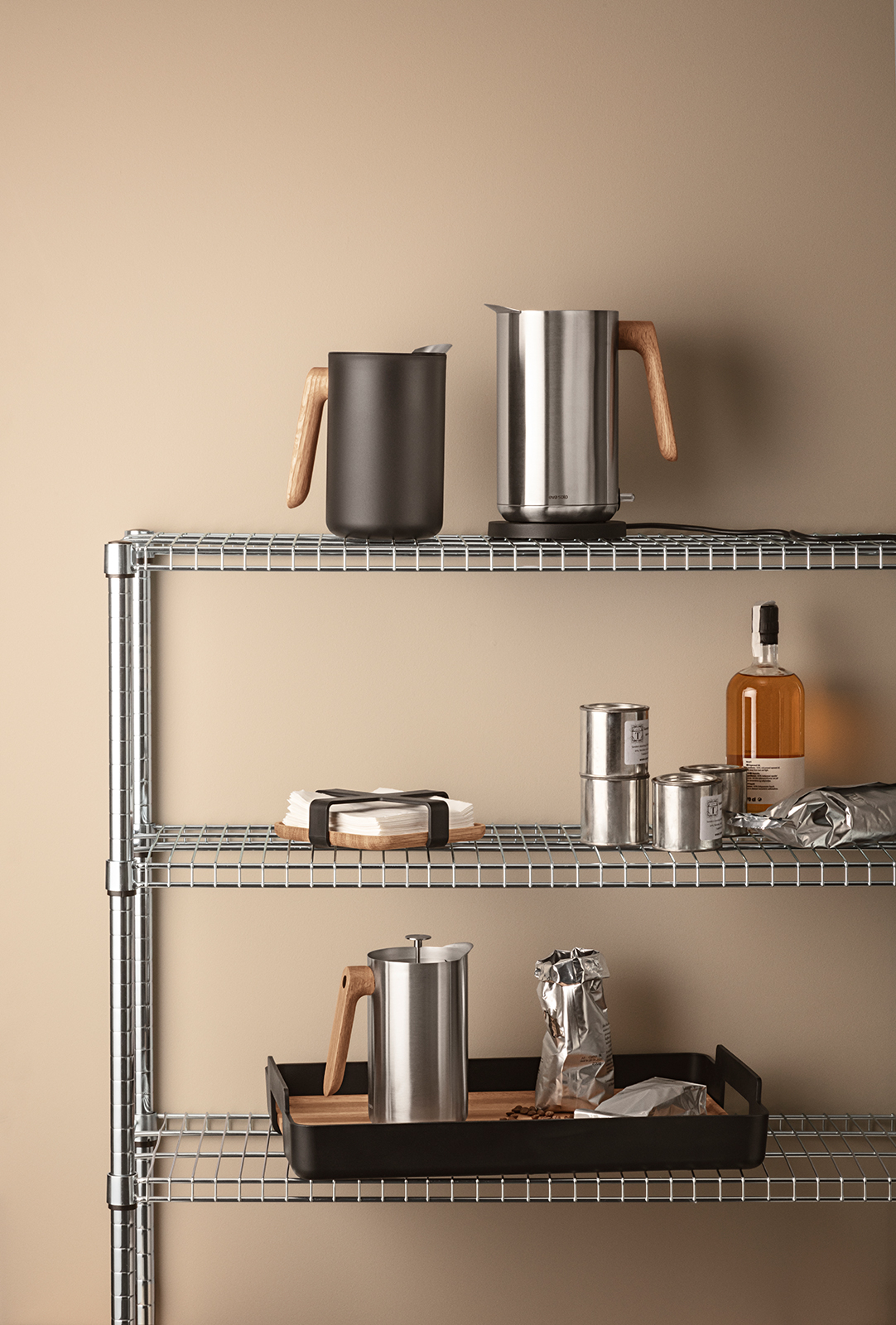 Ein metallregal, das mit verschiedenen Eva Trio Produkten bestückt ist. Auf dem oberen regalbrett steht links die Isolierkanne in schwarz.