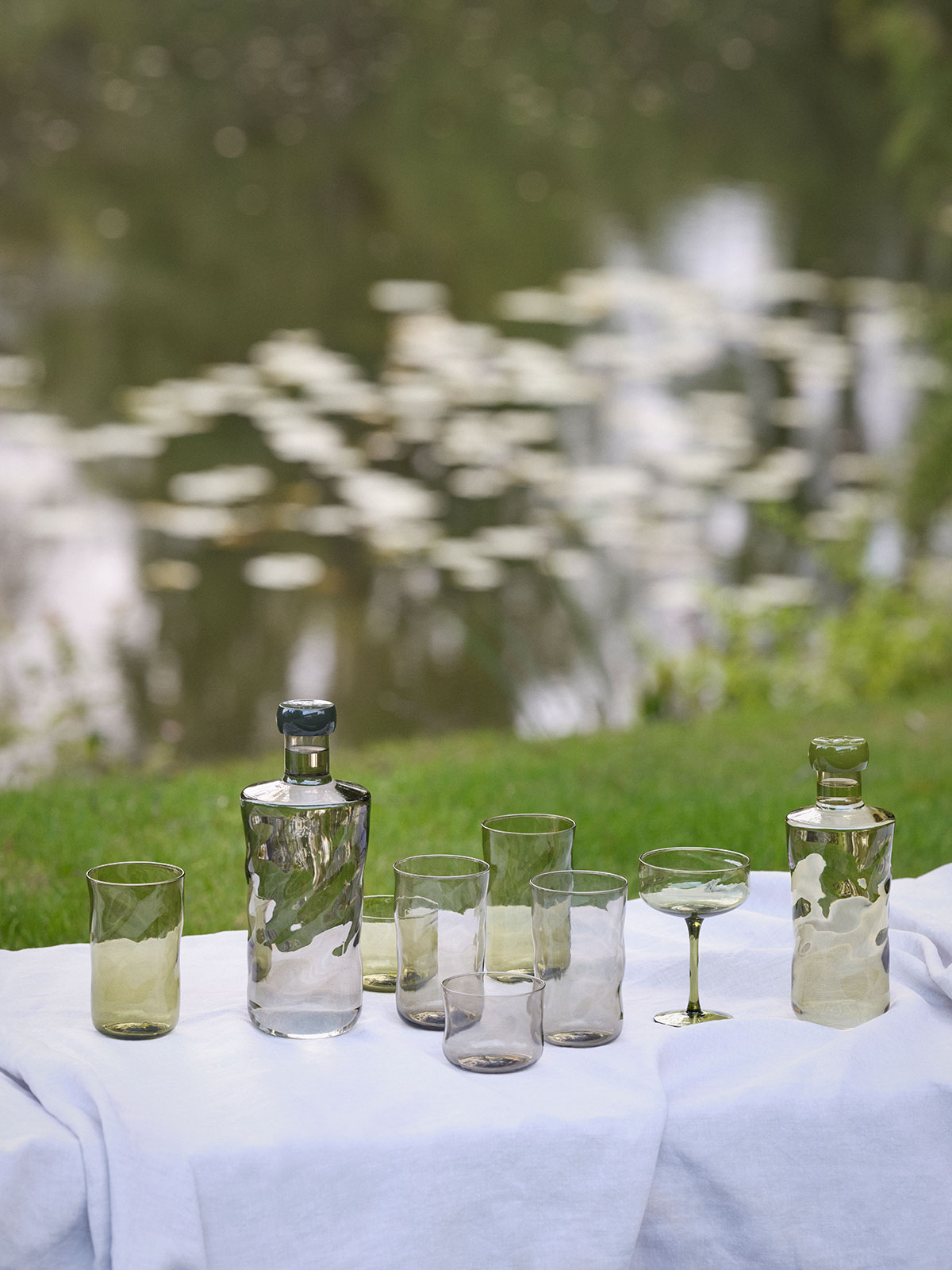Auf einer weißen Tischdecke, ausgebreitet auf grünem Gras, stehen miteinander vermischt Wasserflaschen und Trinkgläser in grün und rauchgrau.