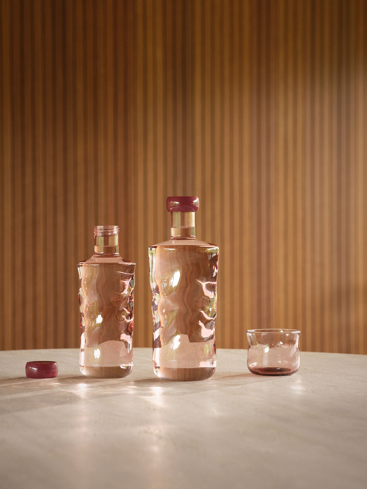 Vor einer holzgetäfelten Wand stehen auf einem Tisch mehrere rote Glasflaschen neben Trinkgläsern der gleichen Farbe.