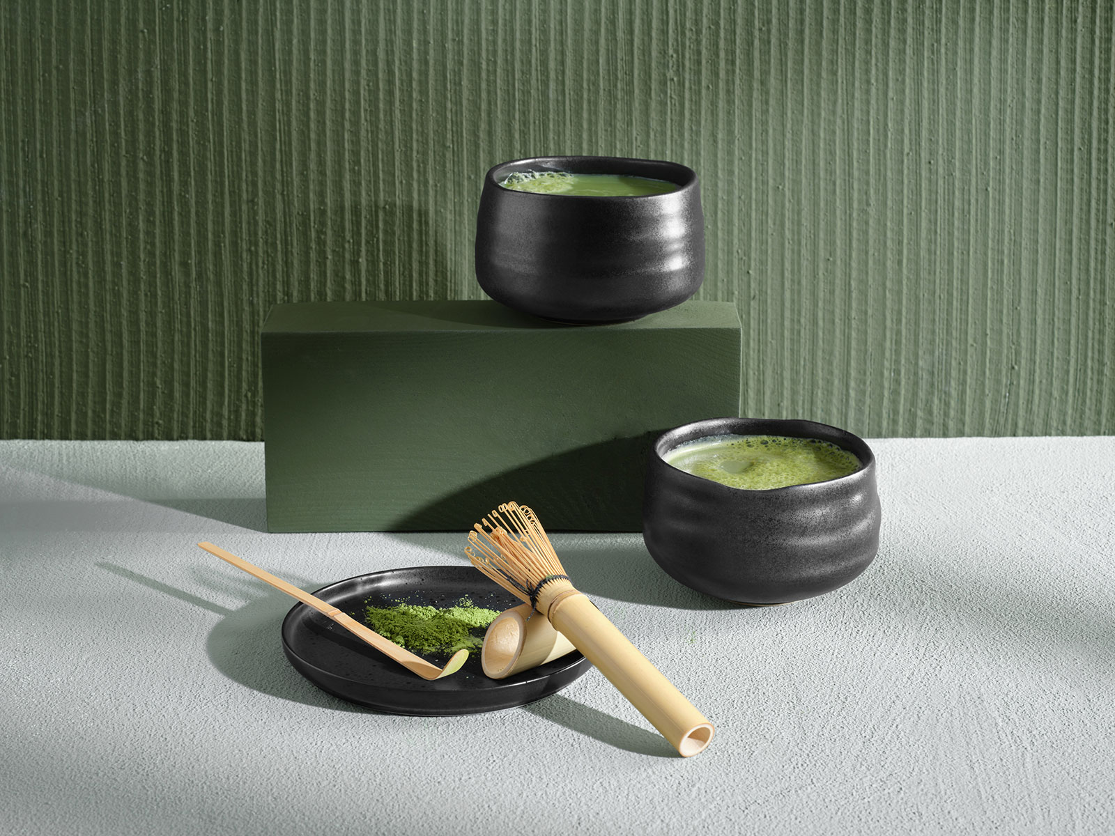 Vor einem grünen Hintergrund stehen zwei Moribana Matcha Schalen mit frisch zubereitetem Matcha. Im Vordergrund liegen auf einem Untersetzer ein Matcha Besen und ein Matcha Löffel.