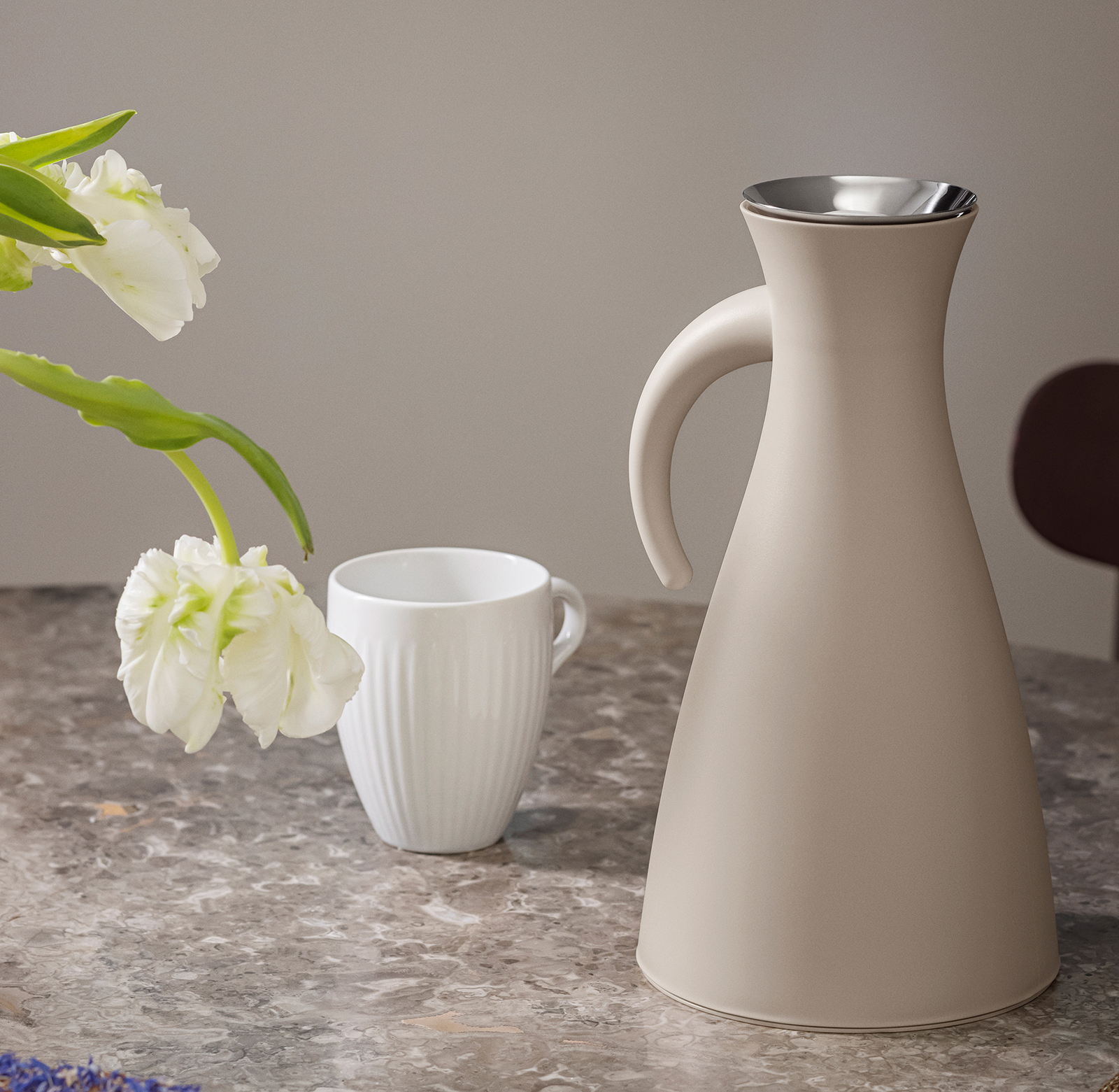 Nahaufnahme eines Tischs, auf dem die Legio Nova Kaffeetasse neben einer Isolierkanne steht. Ins Bild ragt eine Blume.